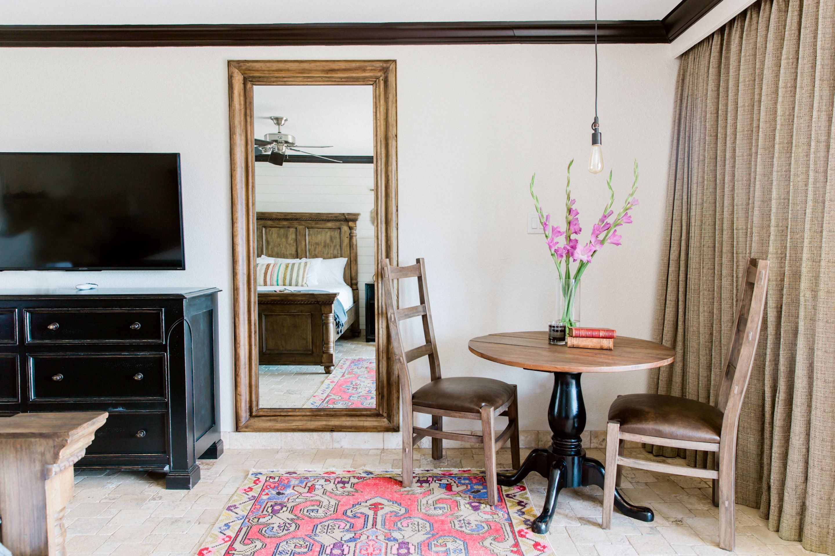 Habitación de hotel con televisión, cómoda de madera, una mesa redonda con sillas, espejo y un arreglo floral.