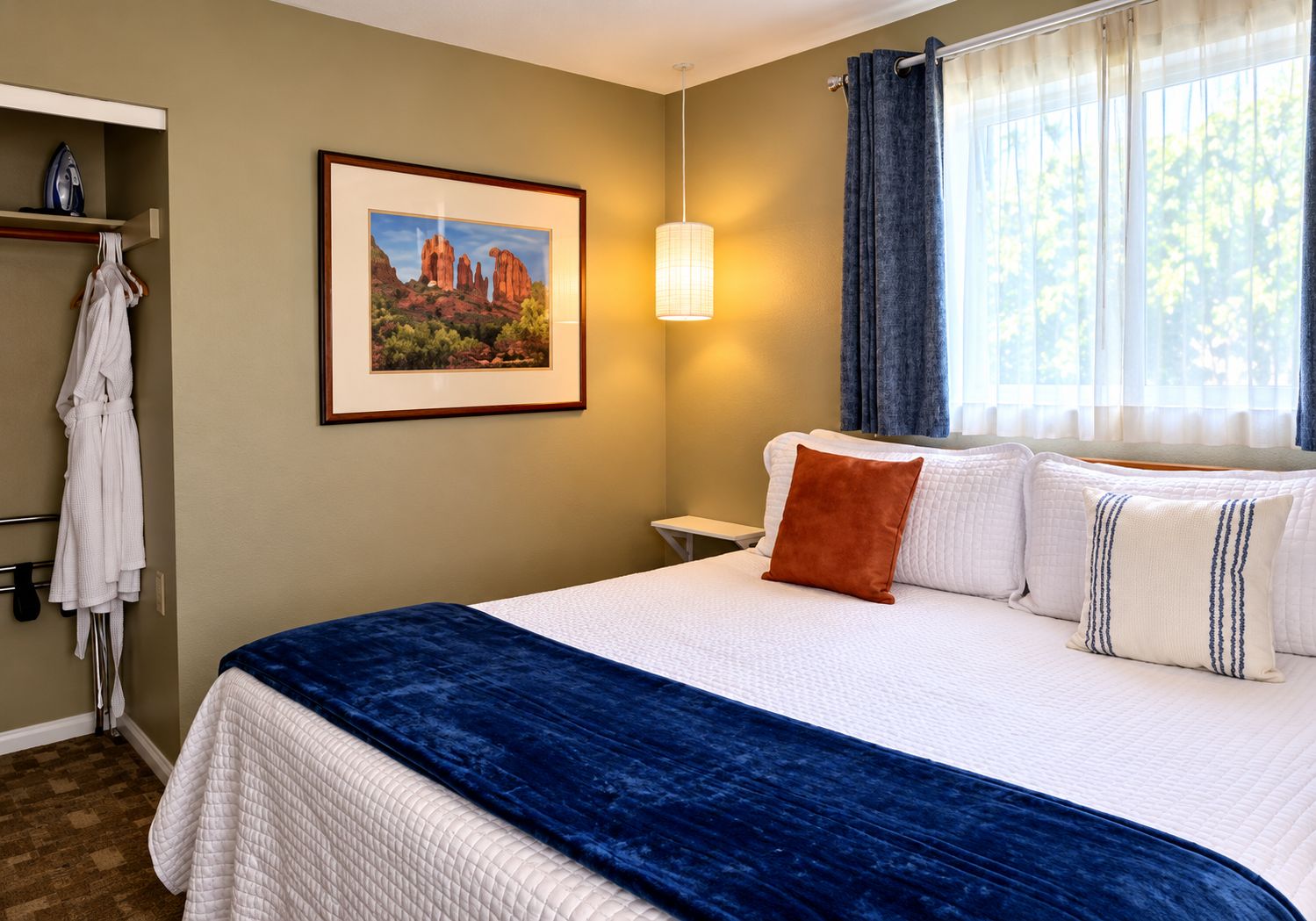 A hotel room with a bed, a painting on the wall, a robe rack, and a window with curtains.