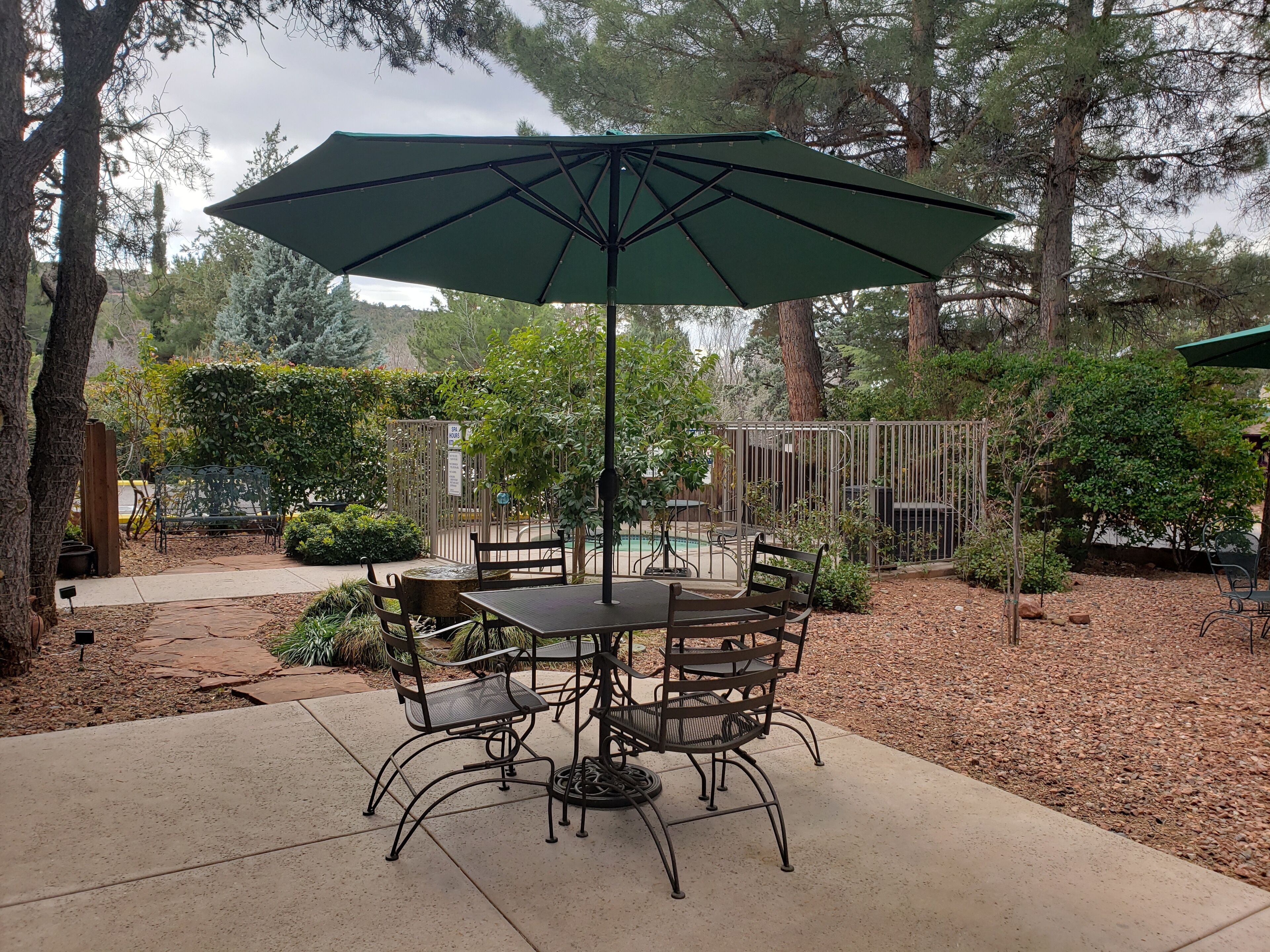 An outdoor patio area with a round table and chairs, a large umbrella, and a pool in the background.