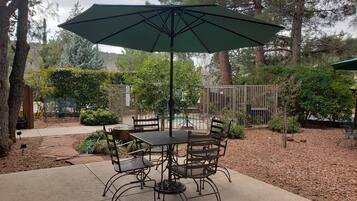 An outdoor patio area with a round table and chairs, a large umbrella, and a pool in the background.