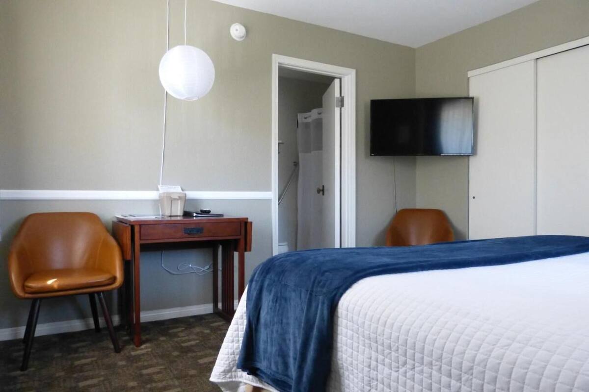 Quarto de hotel com uma cama, uma escrivaninha, duas cadeiras, uma televisão e uma porta que dá para um banheiro.
