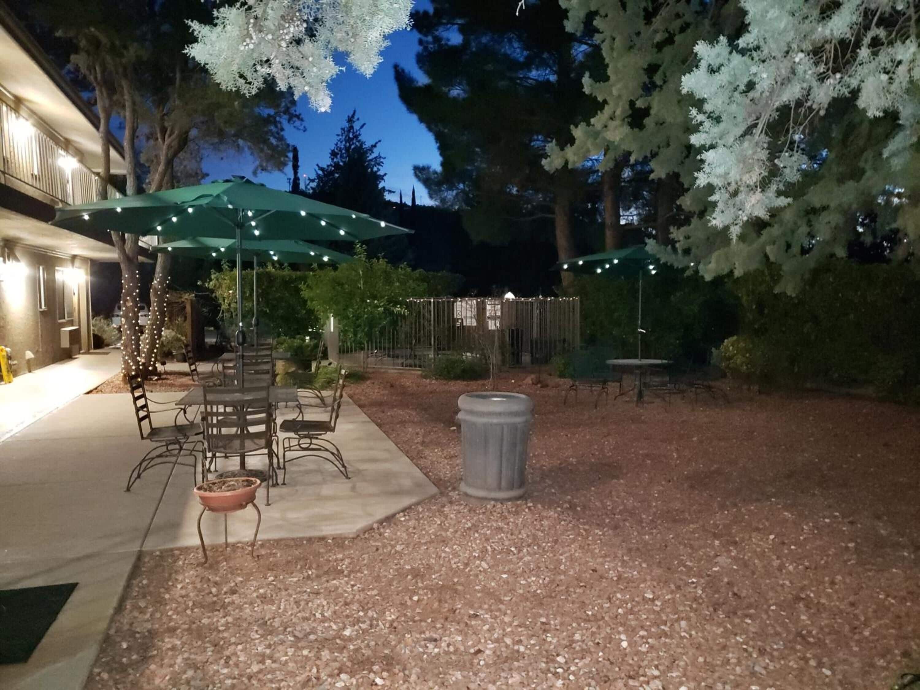 An outdoor patio area with tables and chairs under green umbrellas, surrounded by trees and a gravel path.