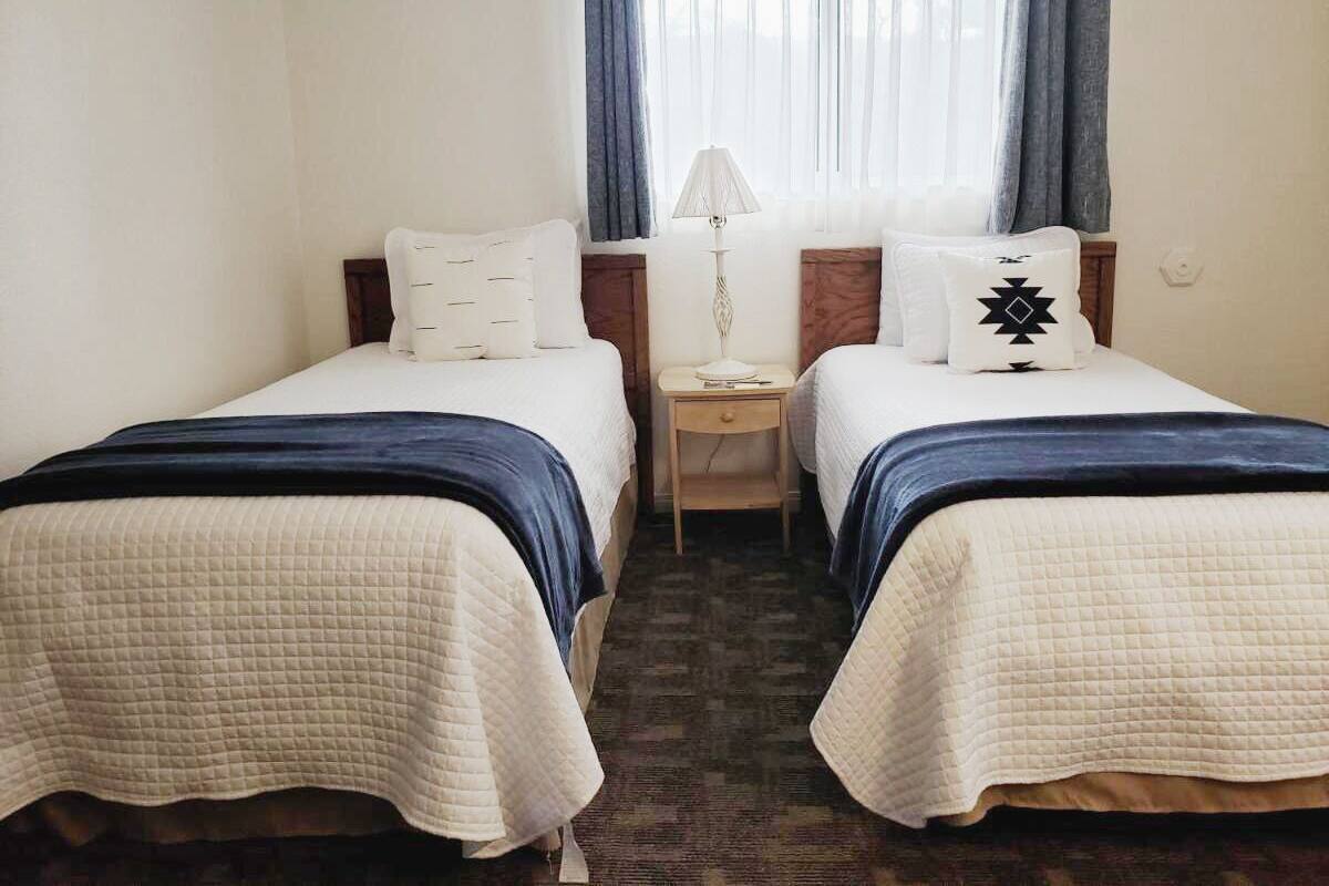 Quarto de hotel com duas camas de solteiro, uma mesinha de cabeceira, uma luminária e uma janela com cortinas.