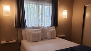 A neatly made bed with a white quilt and two patterned pillows, a bedside table with a lamp, and a window with sheer curtains. - Baby Quail Inn (Sedona)