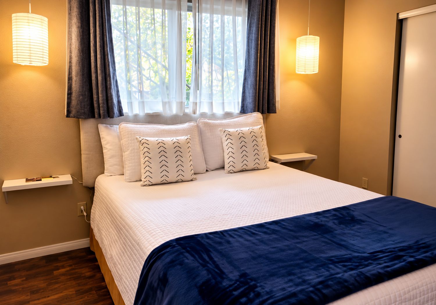 Una habitación de hotel con una cama, dos lámparas de noche, una ventana con cortinas y una puerta.