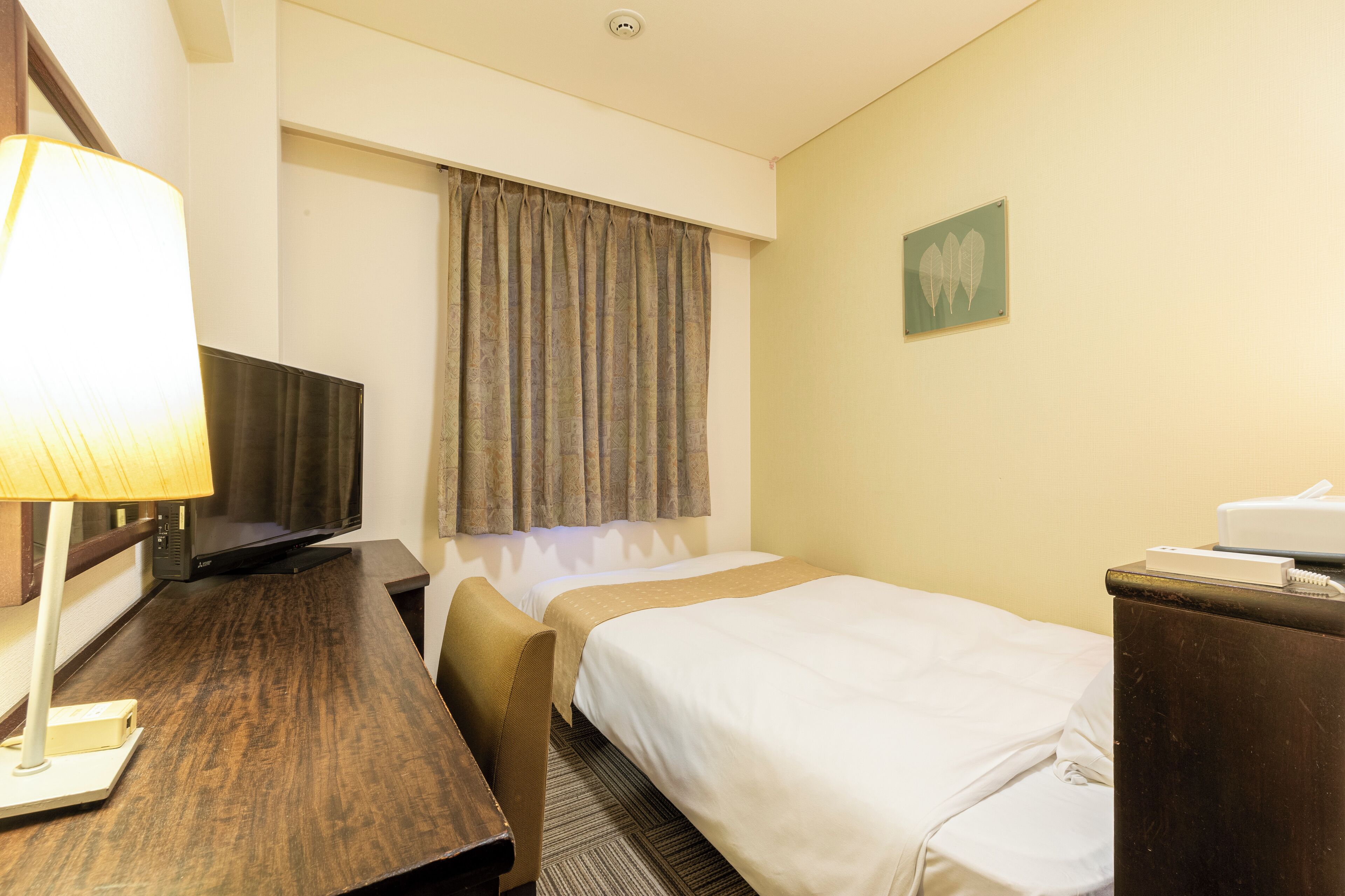 A hotel room with a bed, a desk with a lamp, a television, and a window with curtains.