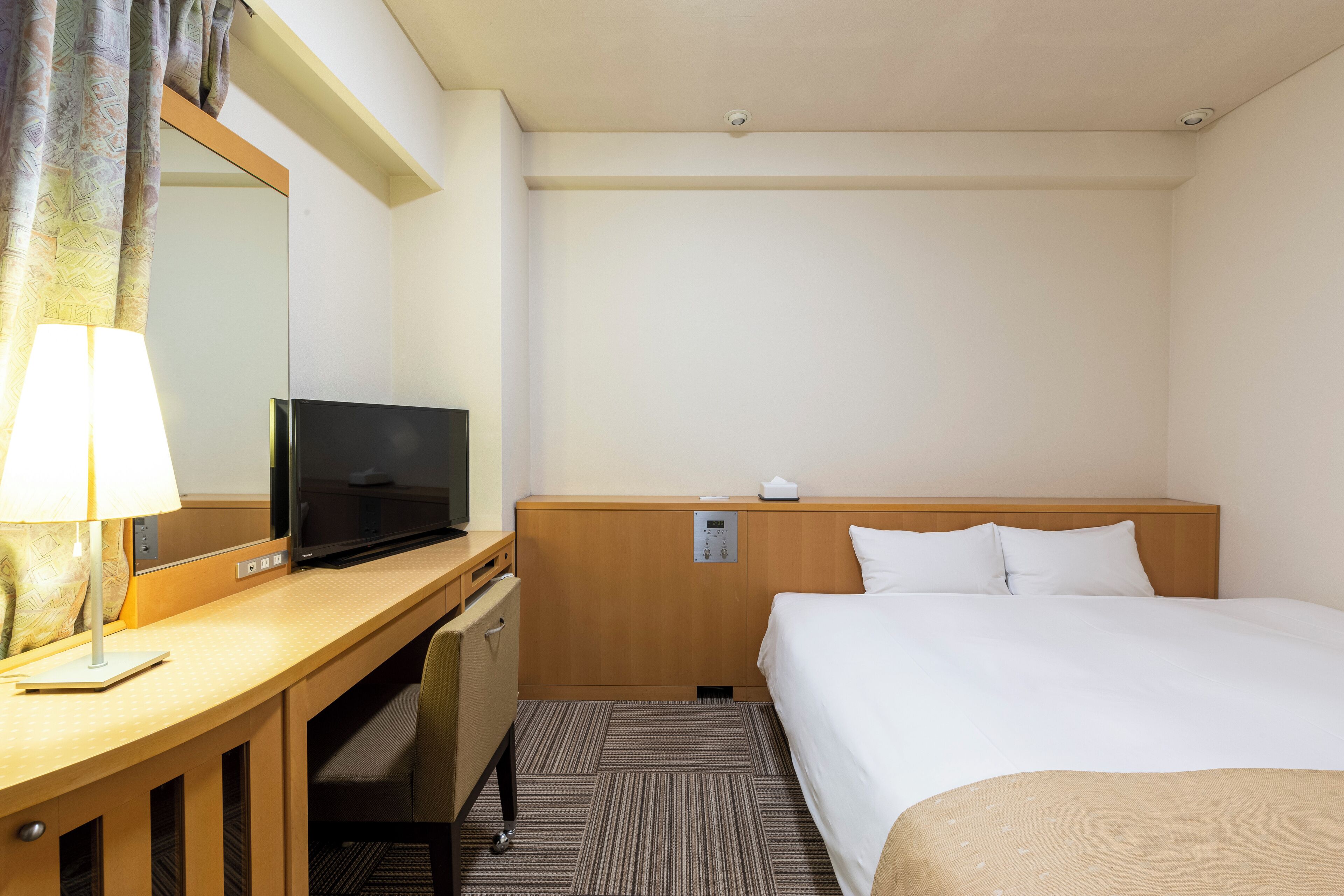 A hotel room with a bed, a desk with a lamp, a television, and a mirror.
