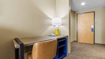 A hotel room with a desk, a lamp, a chair, a door, and a carpeted floor.