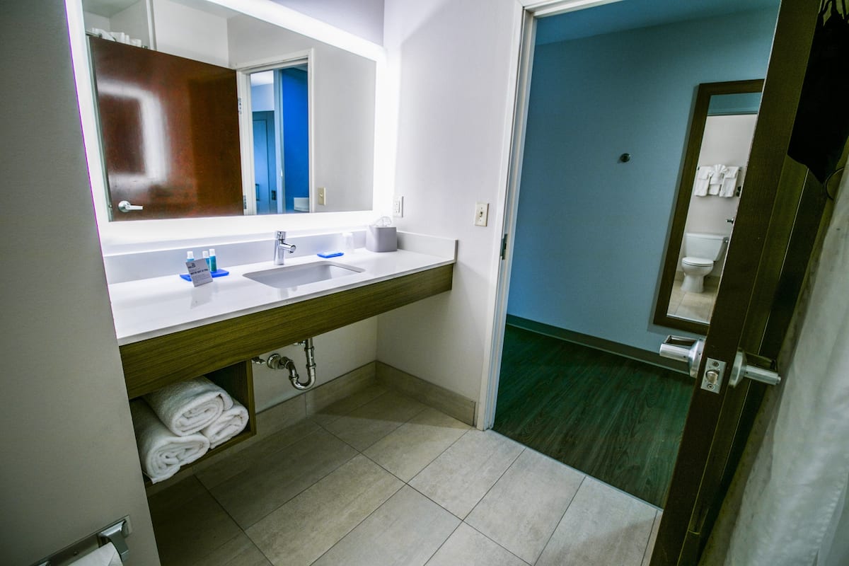 Une salle de bain moderne avec un grand miroir, deux lavabos et des toilettes visibles à travers une porte ouverte.