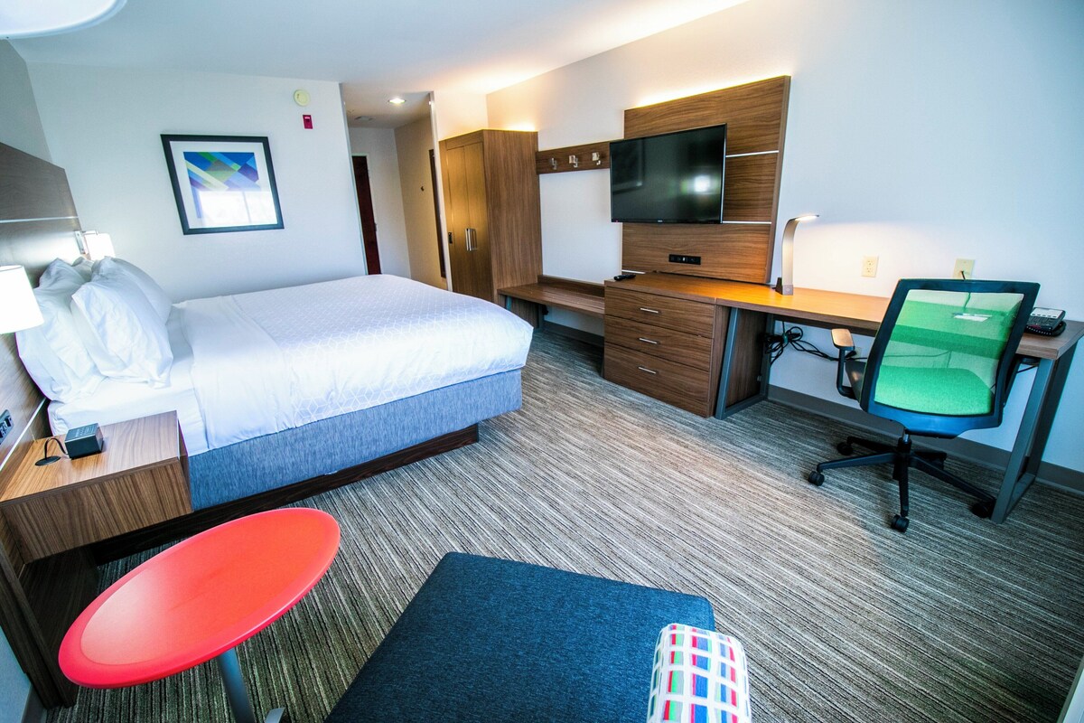 A hotel room with a bed, desk, chair, TV, and a small table with a lamp.