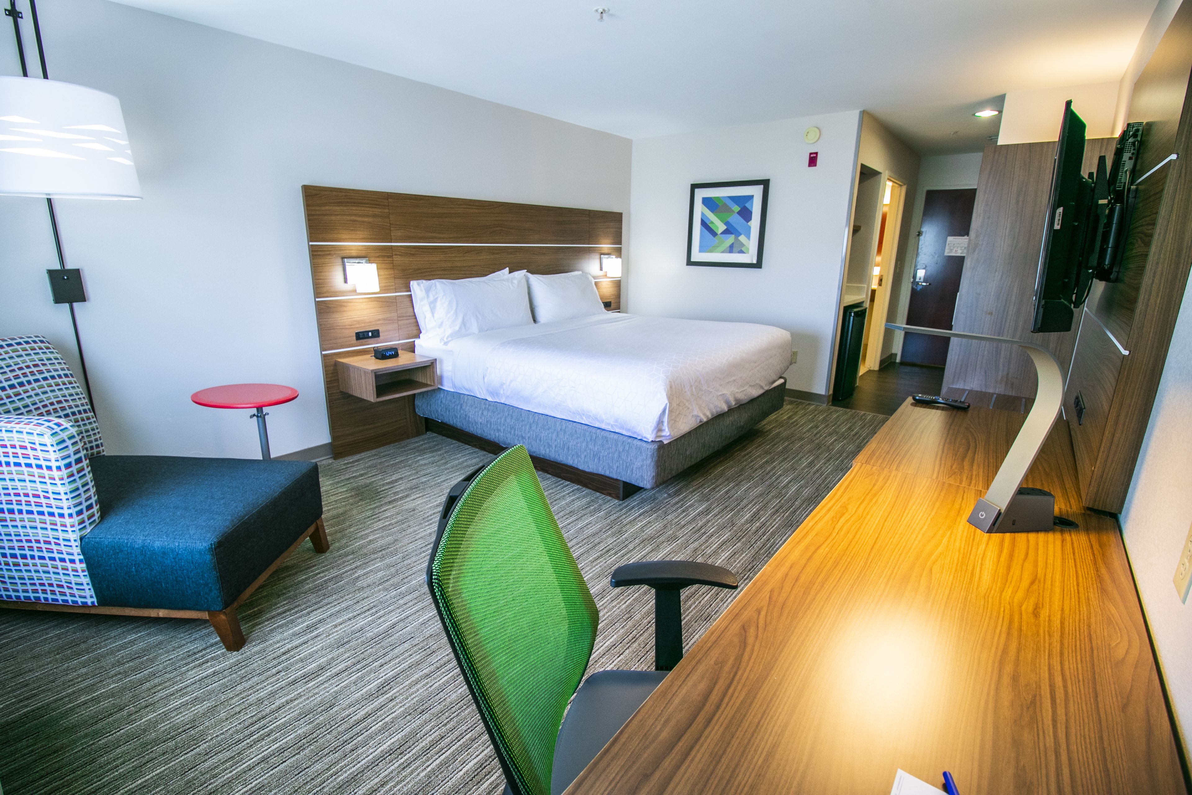 Quarto de hotel com cama, escrivaninha, cadeira, mesa de cabeceira, abajur e uma pequena mesa com base vermelha.