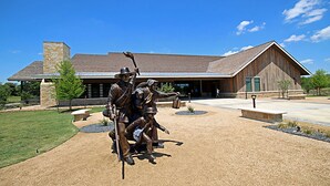 A building with a stone facade, a large wooden barn-like structure, and a statue of people in front. - Holiday Inn Express & Suites Sealy by IHG (Sealy)