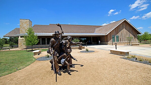 A building with a stone facade, a large wooden barn-like structure, and a statue of people in front. - Holiday Inn Express & Suites Sealy by IHG (Sealy)