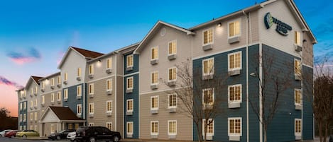 A multi-story hotel with a sign reading "Woodspring Suites." It features a parking lot with several cars and a small tree in front.