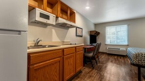A compact kitchen with wooden cabinets, a microwave, and a sink. There's a dining area with a table and chairs, and a bed with a patterned cover. - WoodSpring Suites Macon North I-75 (Macon)