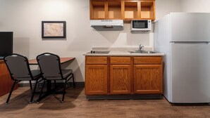 A compact kitchen with wooden cabinets, a white refrigerator, a microwave, a sink, and two chairs. - WoodSpring Suites Macon North I-75 (Macon)