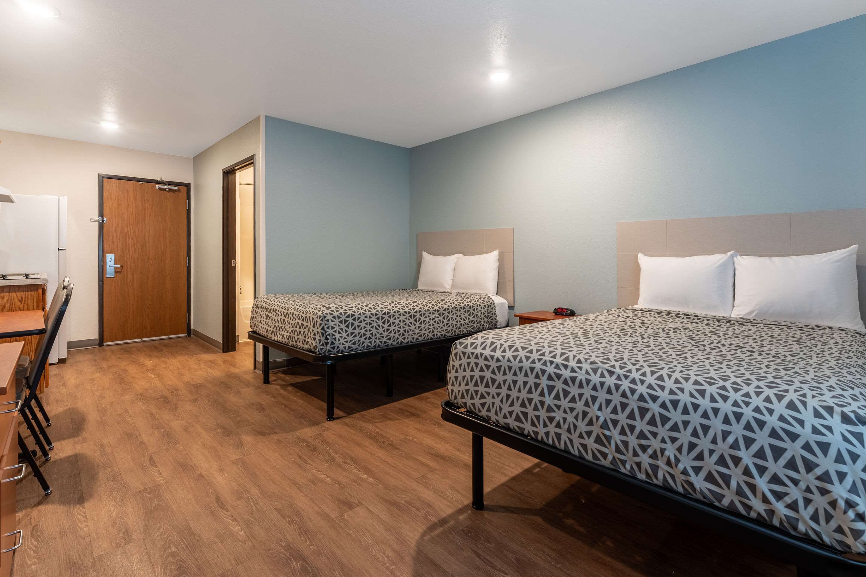 A hotel room with two beds, a wooden floor, a desk with a chair, and a door.