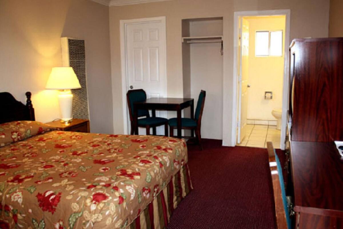 A hotel room with a bed, a desk, and a chair. There is a lamp on the nightstand and a wardrobe in the corner.