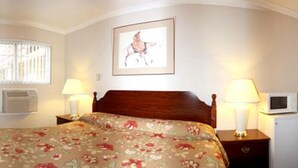 A hotel room with a wooden bed, floral bedspread, two bedside lamps, a framed picture on the wall, a microwave, and a small table with a lamp. - Sunset Inn and Suites (West Sacramento)