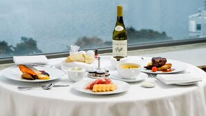 A table set with various dishes and a bottle of wine, overlooking a body of water and high-rise buildings.