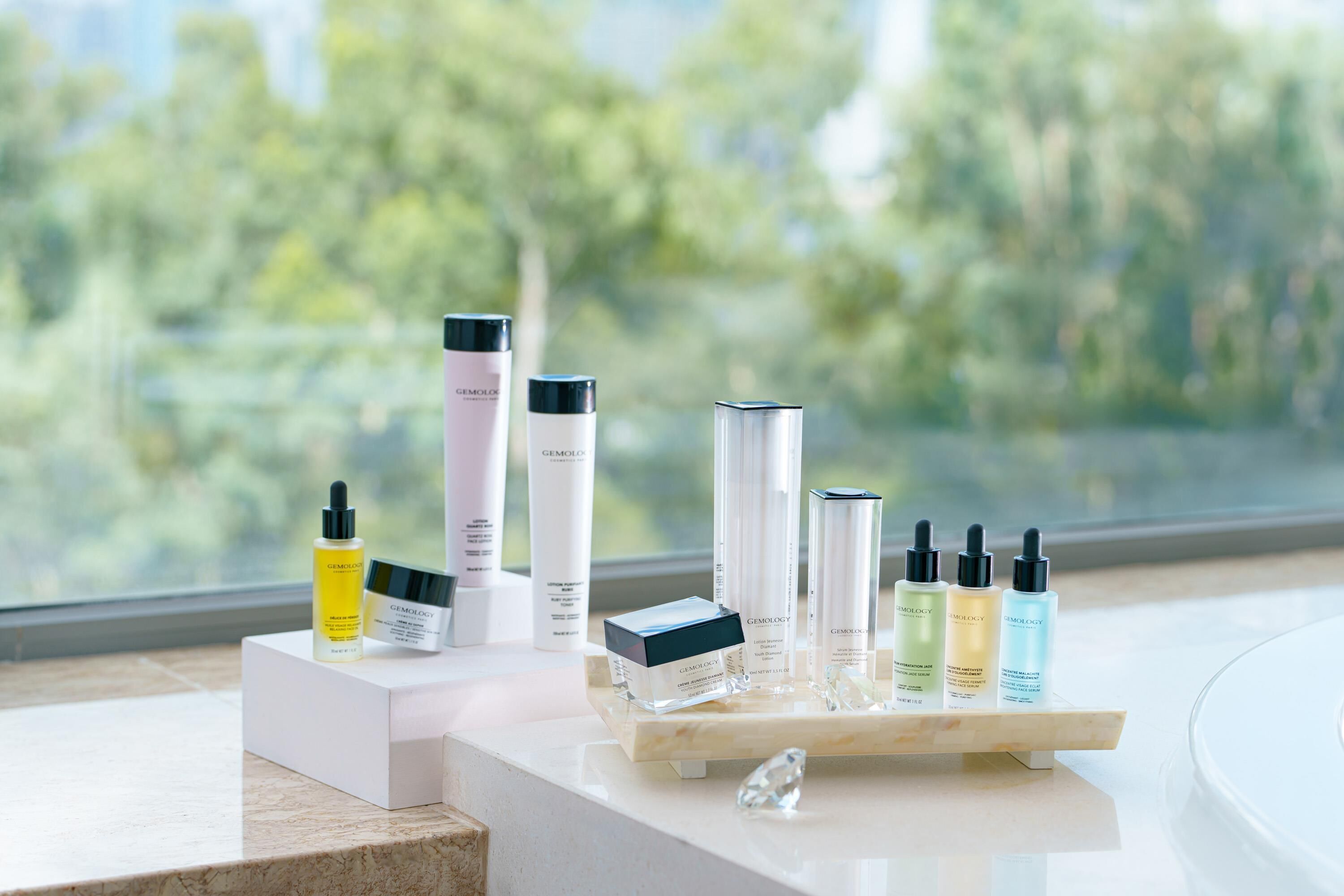A display of skincare products on a marble countertop with a view of trees through a window.