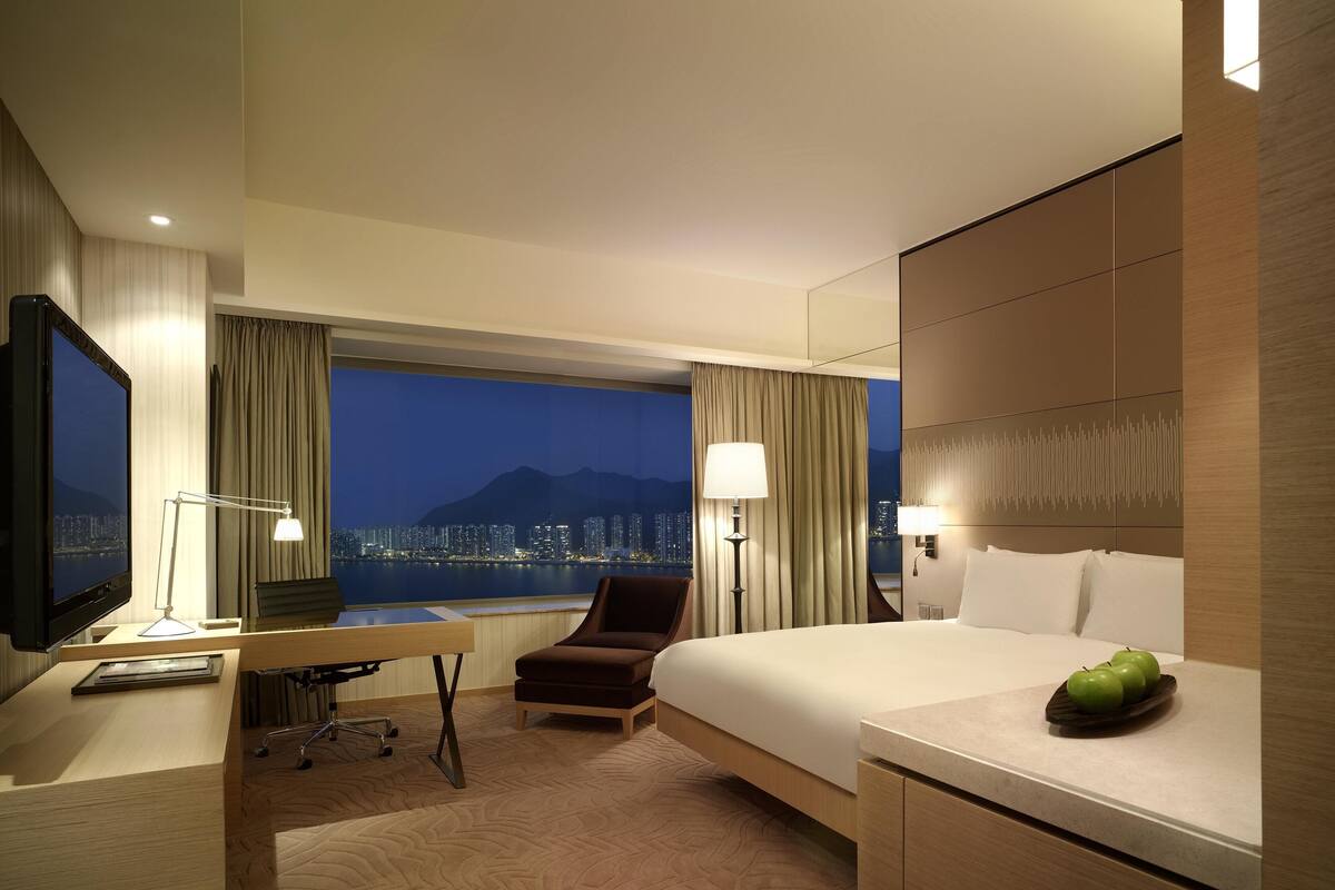 A hotel room with a large bed, a desk, a chair, a TV, and a view of a cityscape at night.