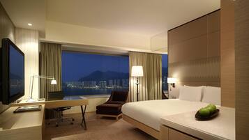 A hotel room with a large bed, a desk, a chair, a TV, and a view of a cityscape at night.