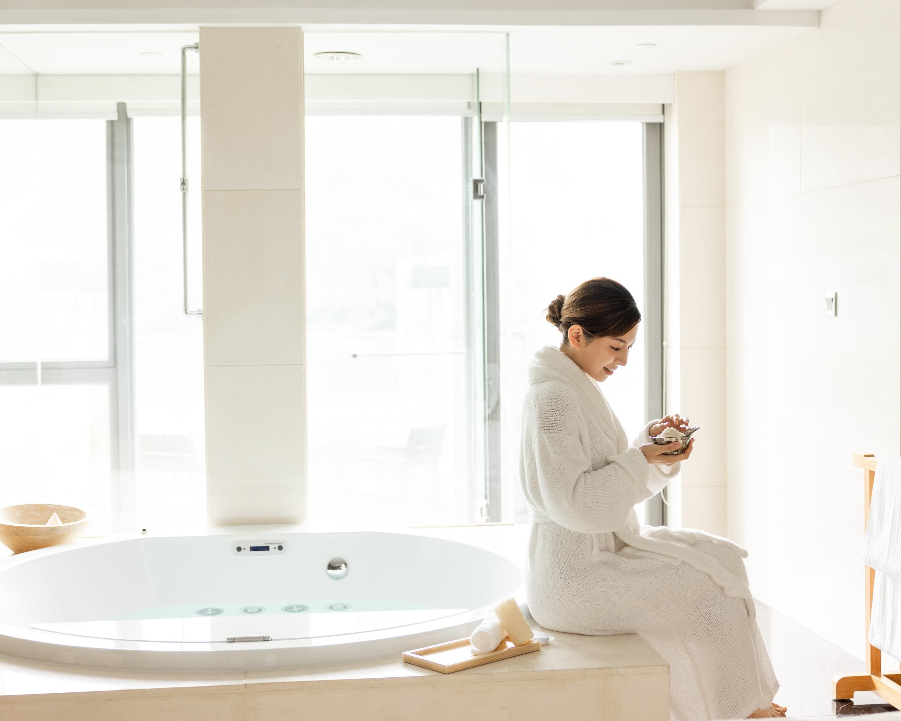 A person in a white bathrobe sitting by a large bathtub with a wooden stool, in a bright room with large windows.