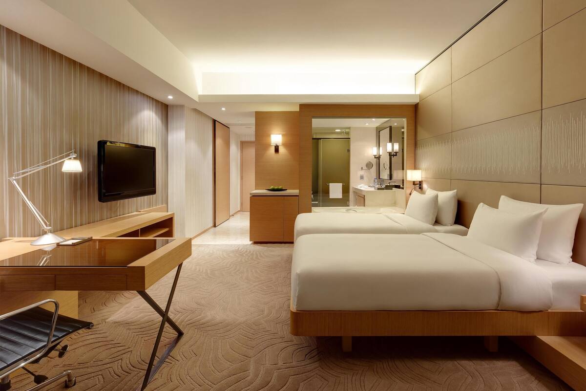 A hotel room with a bed, bedside tables, a TV, a desk with a lamp, a bathroom with a mirror, and a shower area.