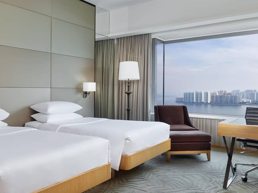 Ein Hotelzimmer mit zwei Betten, einem Schreibtisch, einem Stuhl, einer Lampe und Blick auf eine Stadtlandschaft.