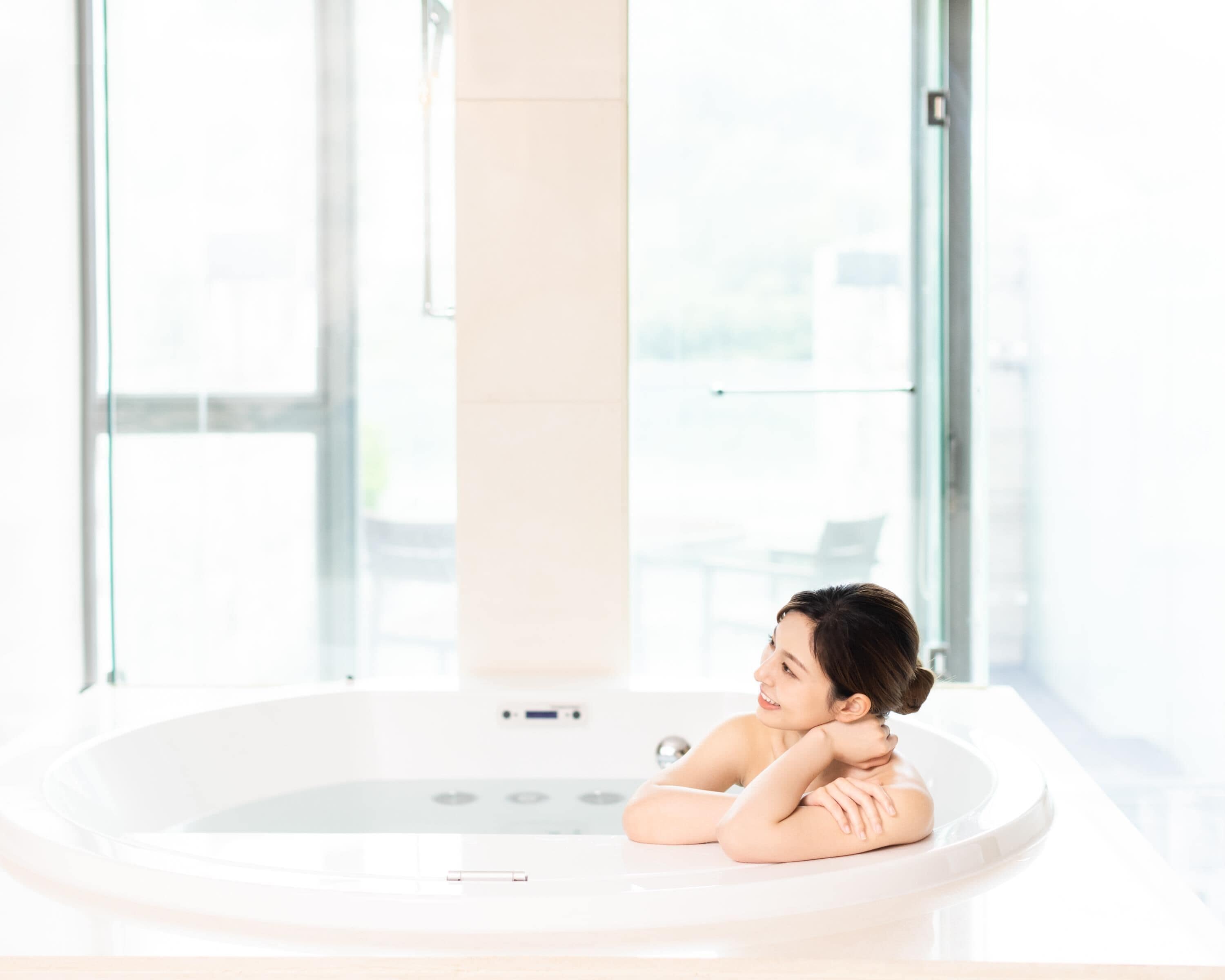 Eine Person entspannt sich in einem modernen, hell einfallenden Badezimmer mit großem Fenster, einer Badewanne und einem Waschbecken.