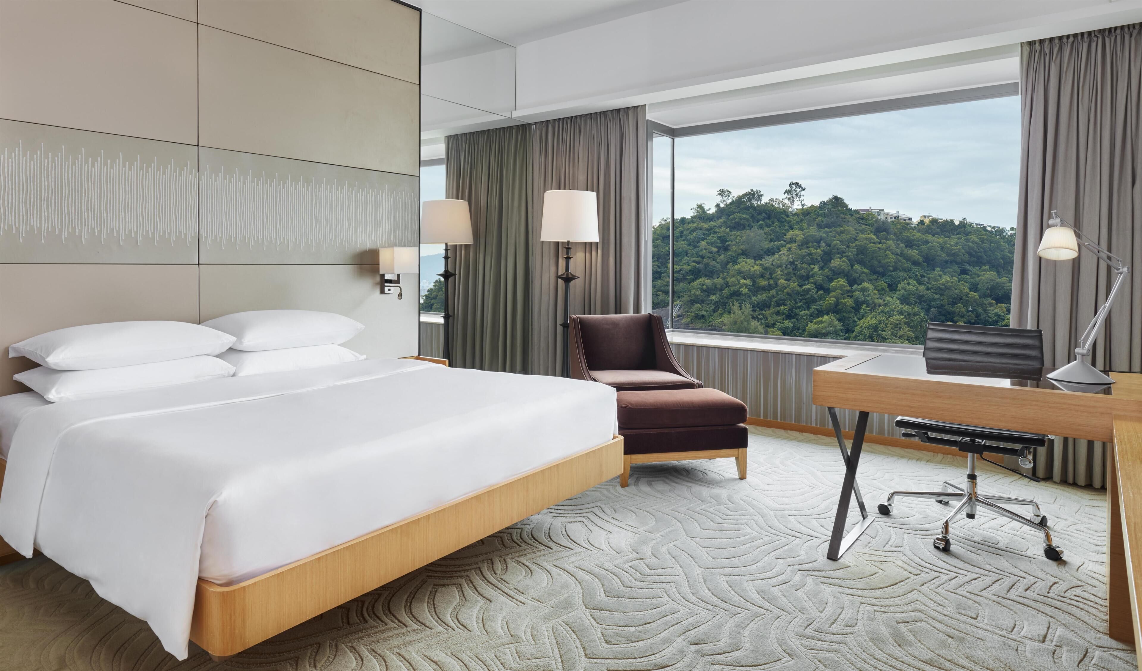 A hotel room with a large bed, a desk, a chair, and a view of a forested area.
