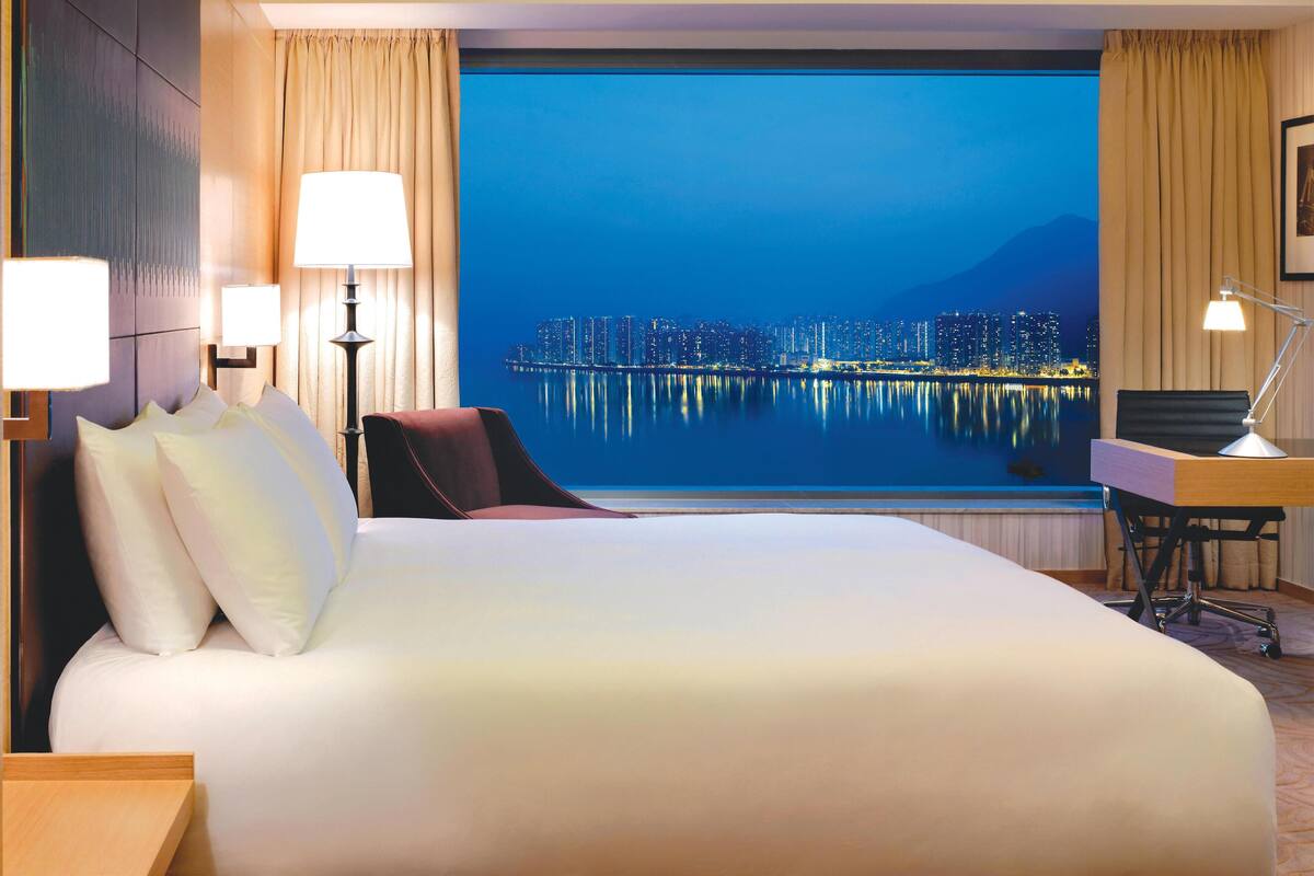 A hotel room with a large bed, a desk with a chair, and a view of a cityscape at night.