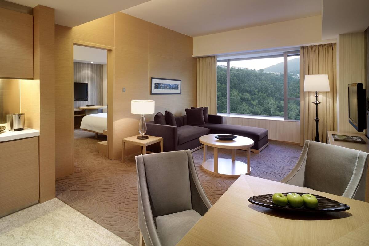 A hotel room with a sofa, a dining table with apples, a TV, and a large window with a view of the forest.