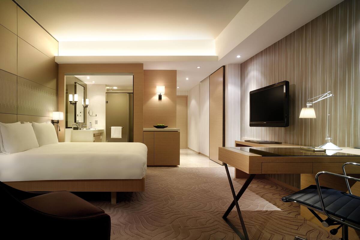 A modern hotel room with a bed, a desk, a TV, and a bathroom visible through an open door.