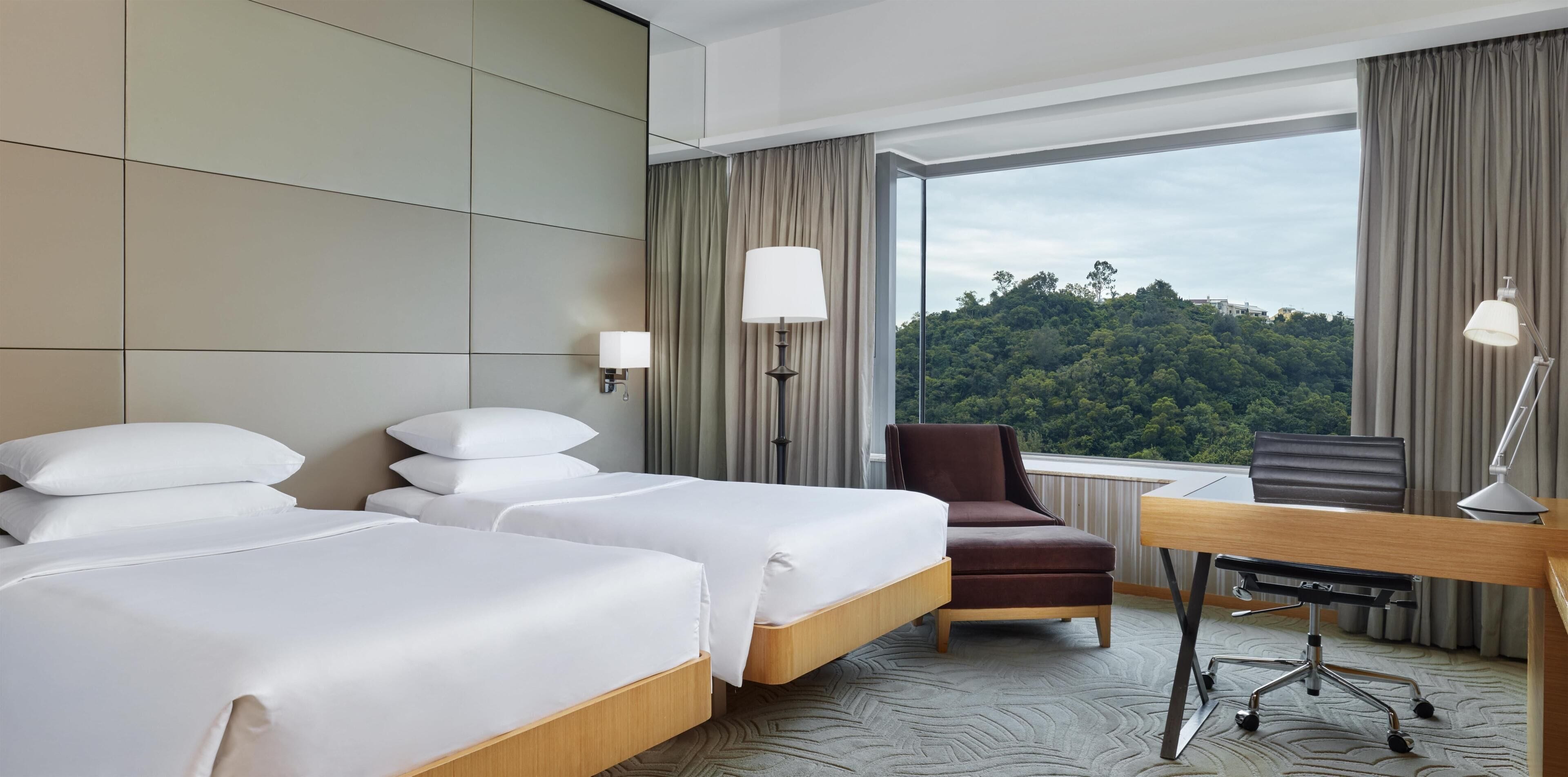 A hotel room with two beds, a desk, a chair, and a large window offering a view of a forested area.