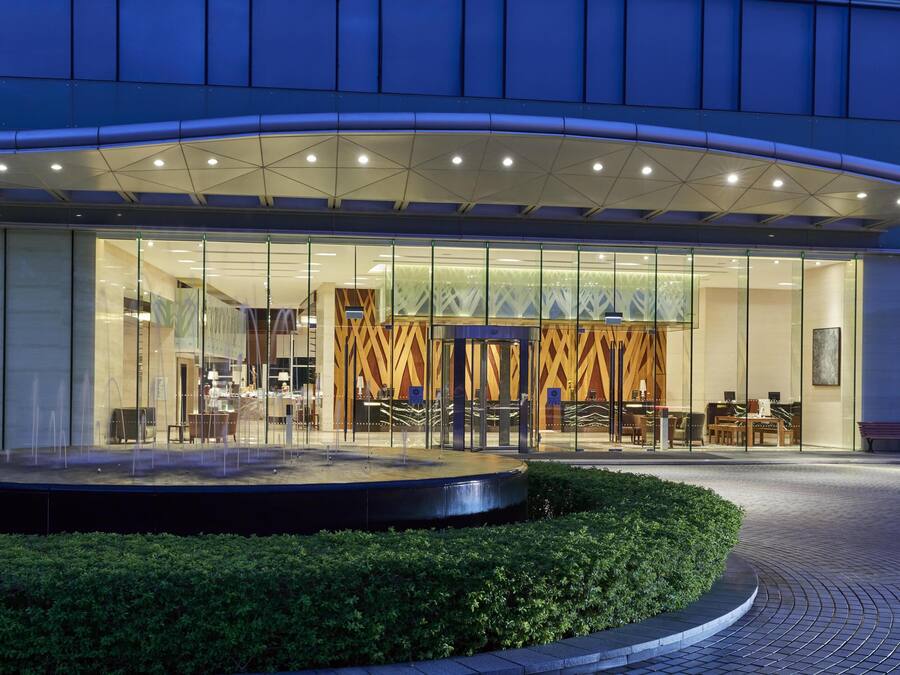 A modern hotel entrance with a glass facade, a reception desk, and a seating area visible through the glass.