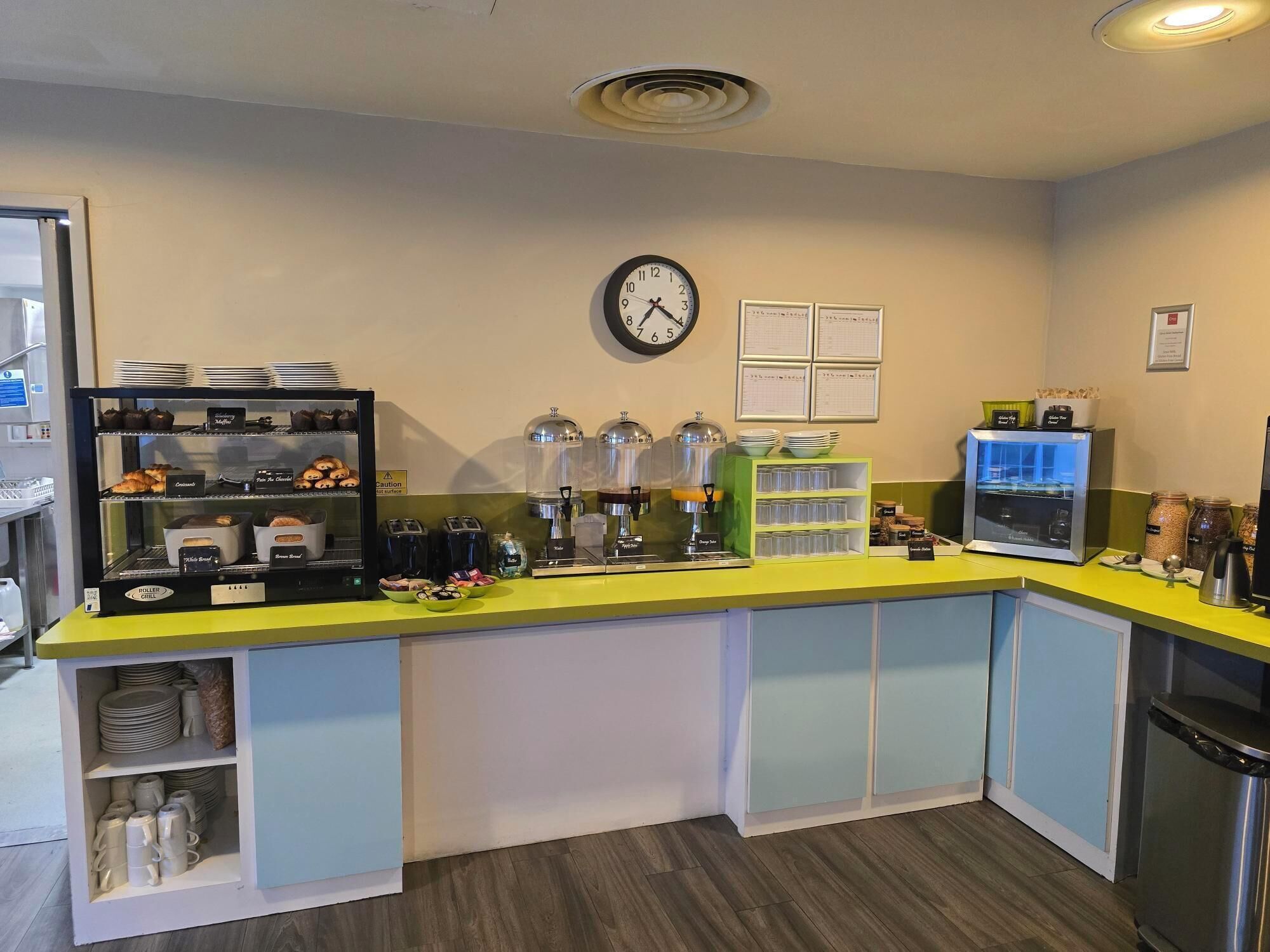 A breakfast bar with a clock, coffee machines, and a display of pastries.
