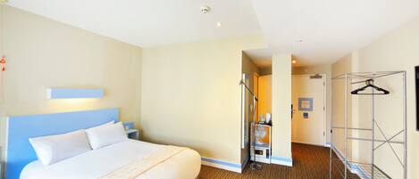 A hotel room with a bed, a bedside table, a wall-mounted light, a shower area, and a coat rack.