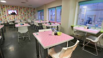 A dining area with tables and chairs, a television mounted on the wall, and windows with curtains.