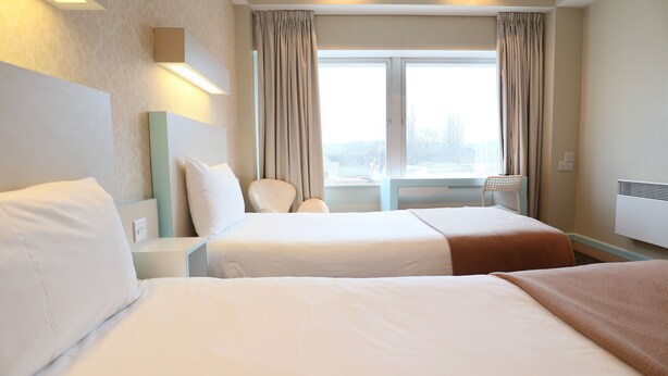 A hotel room with two beds, a window with curtains, a chair, and a wall-mounted heater. - Citrus Hotel Cheltenham by Compass Hospitality (Cheltenham)