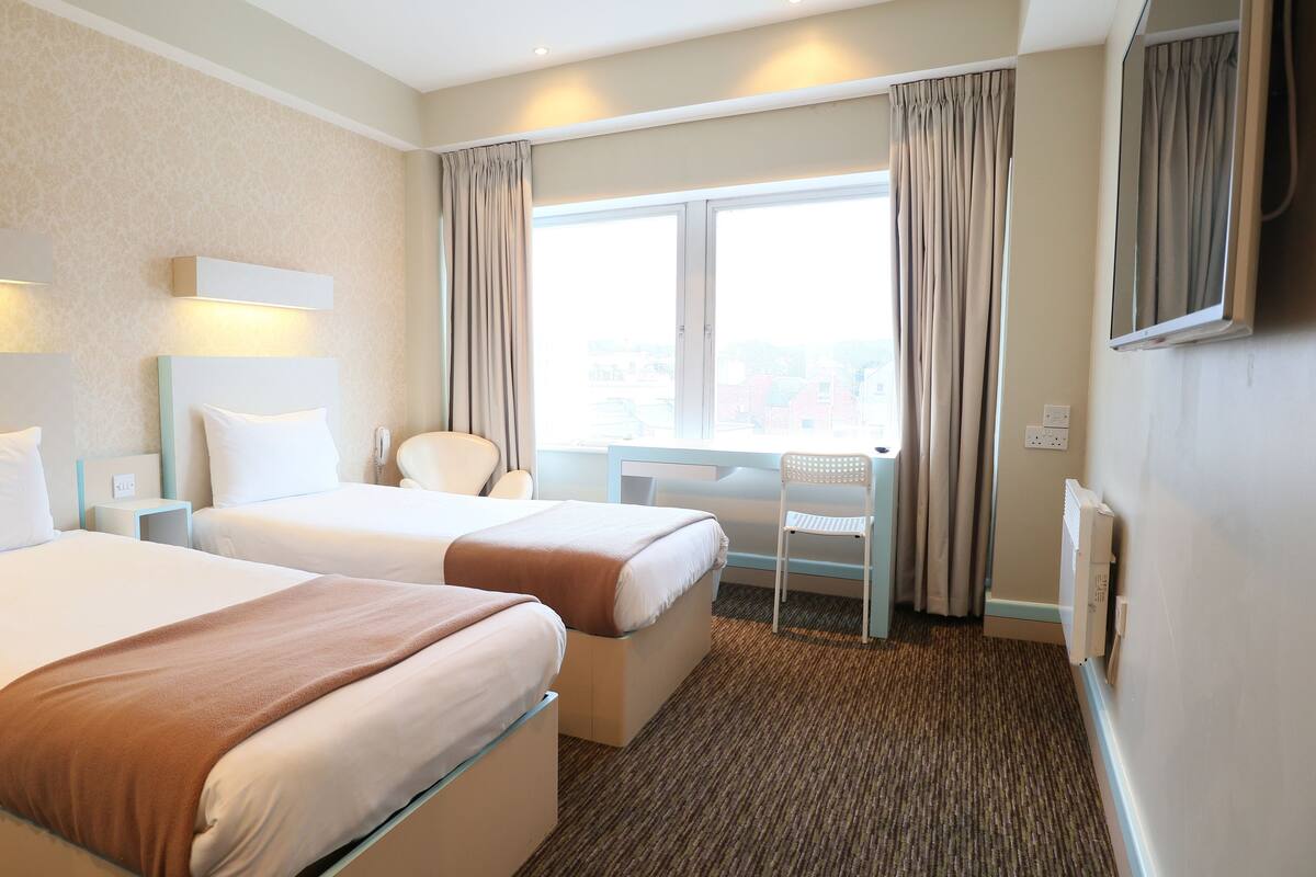 A hotel room with two beds, a desk, a chair, a large window with curtains, and a flat-screen TV mounted on the wall.