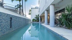 Una piscina con borde de azulejos, rodeada por un edificio blanco con columnas, palmeras y un cielo despejado.
