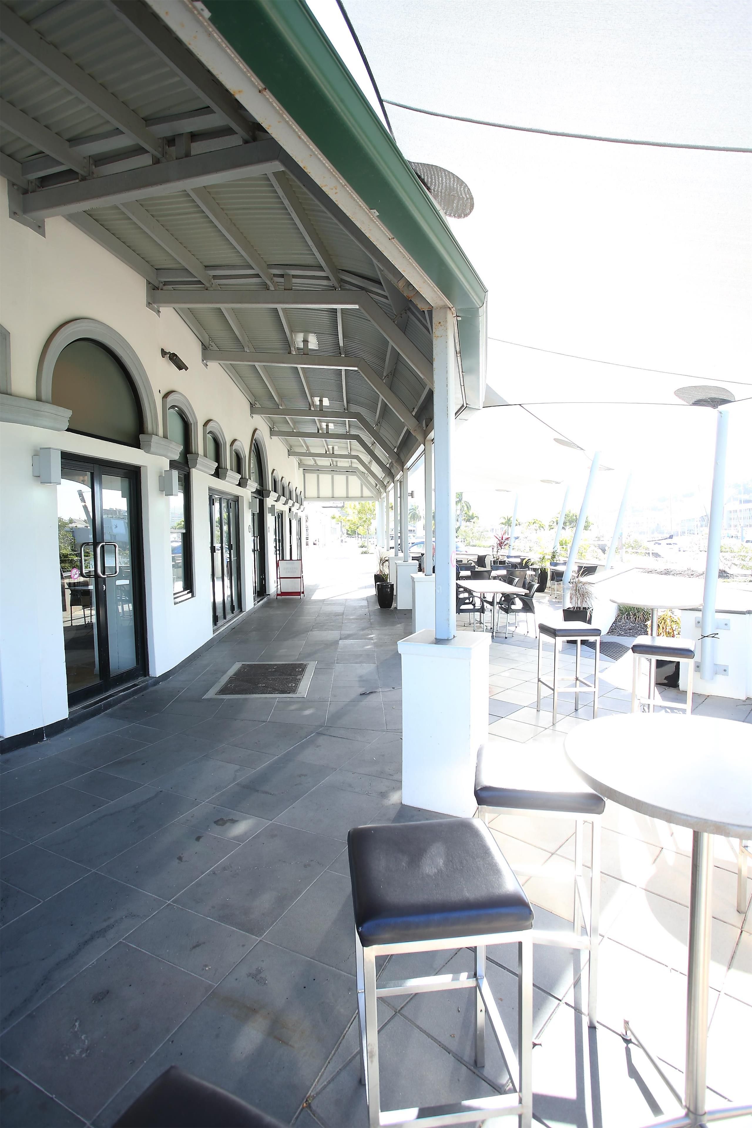 A covered outdoor seating area with tables and chairs, a tiled floor, and a building with arched windows and a green roof.
