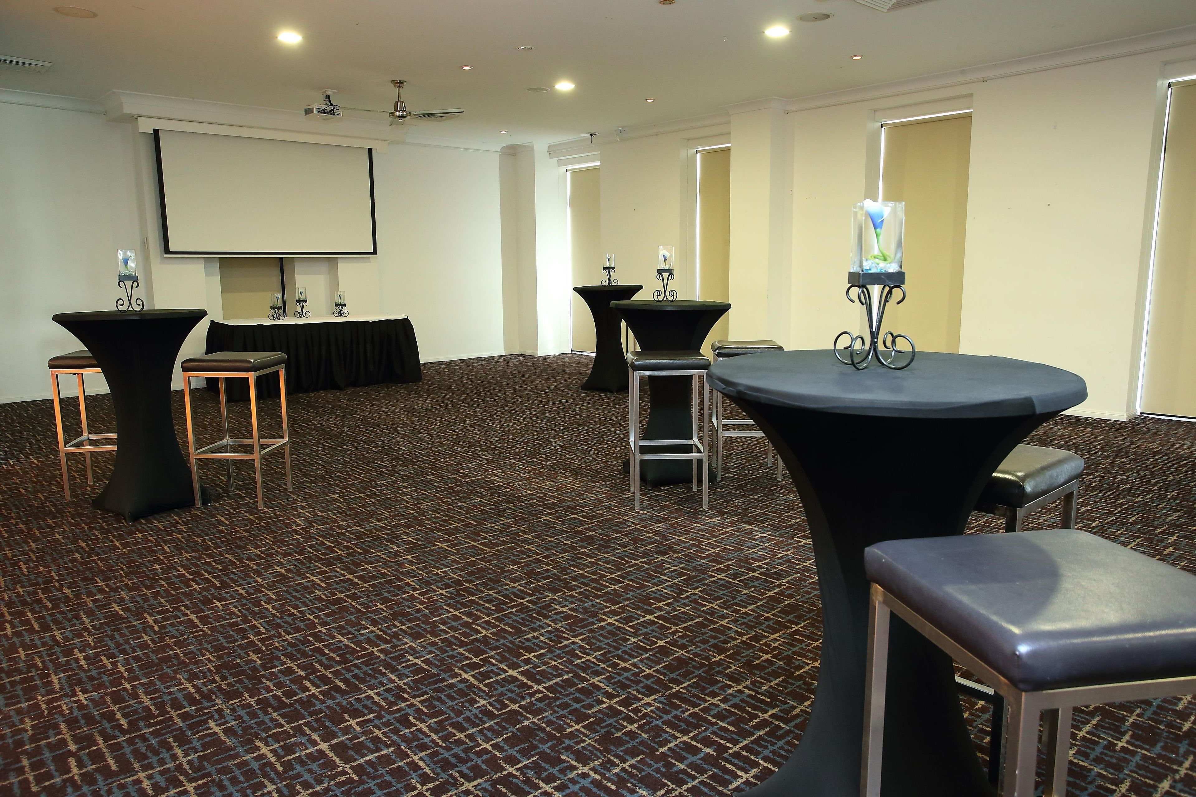 Sala de conferências com várias mesas e cadeiras, uma tela de projetor e piso coberto por carpete.
