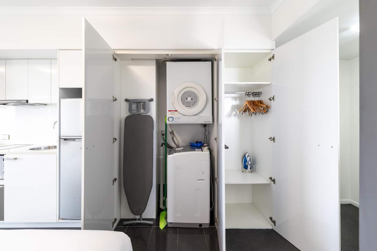 A compact room with a built-in washer and dryer, a small ironing board, and a shelf with hanging clothes.