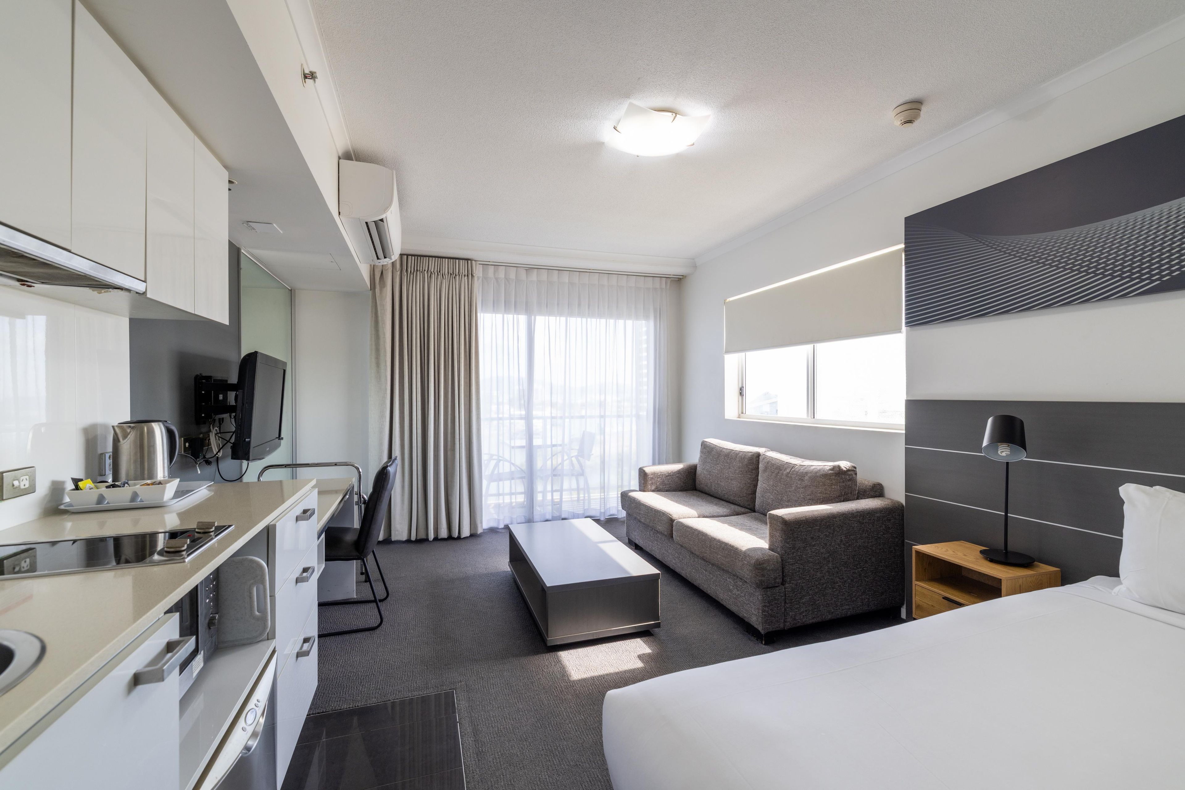 A modern hotel room with a kitchenette, a sofa, a bed, and a large window.