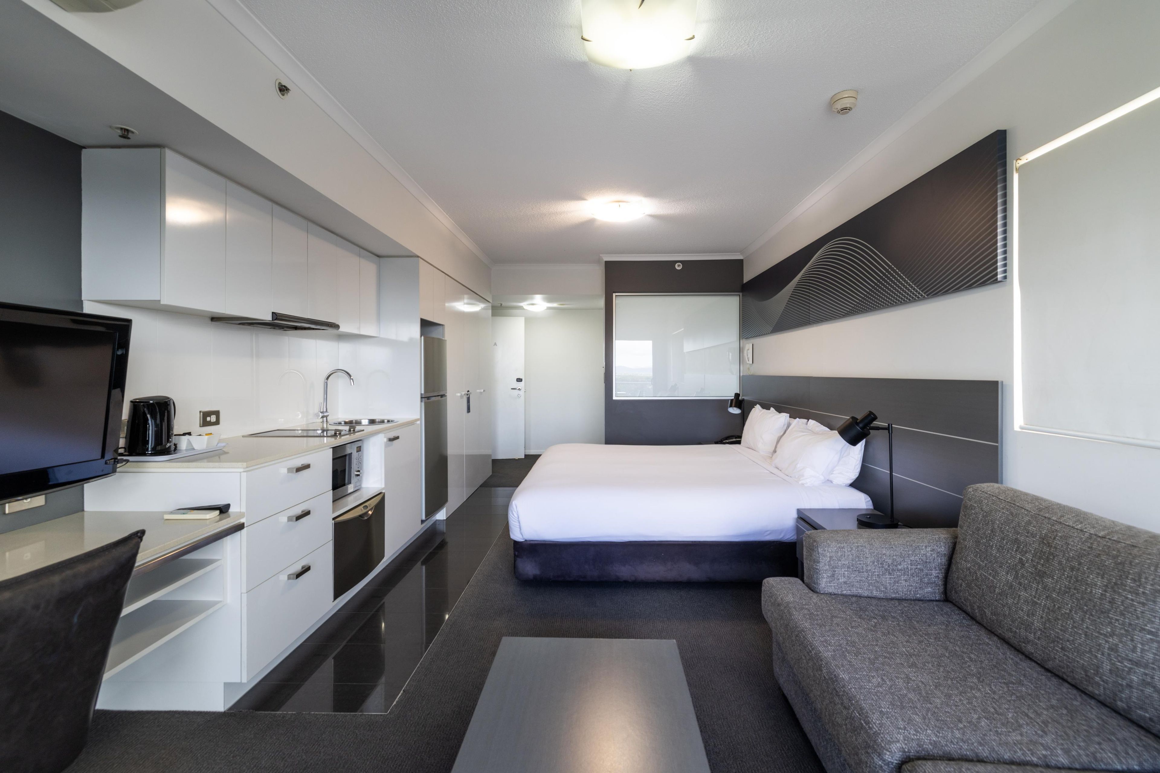 A modern hotel room with a kitchenette, a bed, a sofa, and a flat-screen TV.