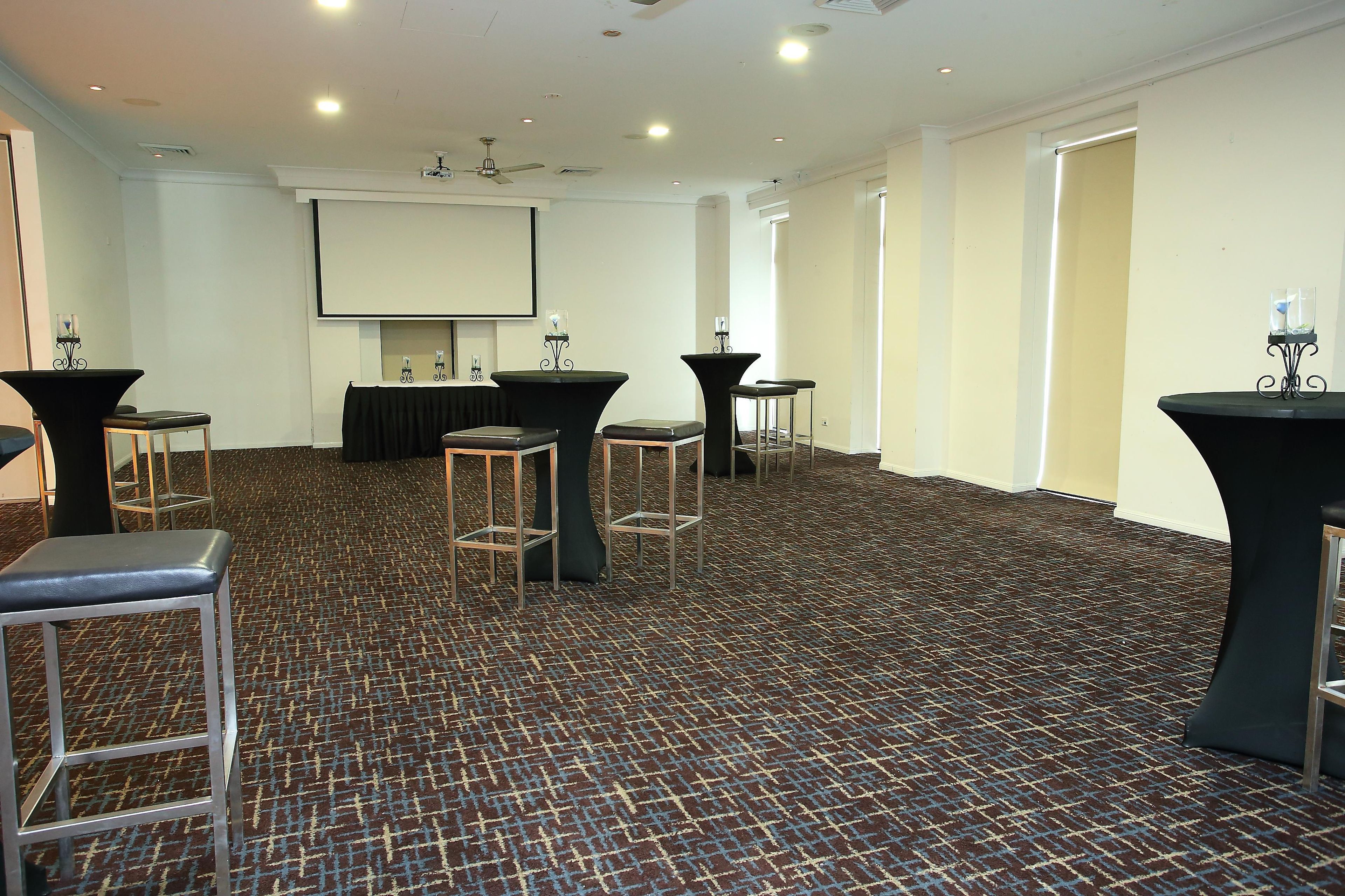 A conference room with a carpeted floor, tables with stools, a projector screen, and ceiling fans.