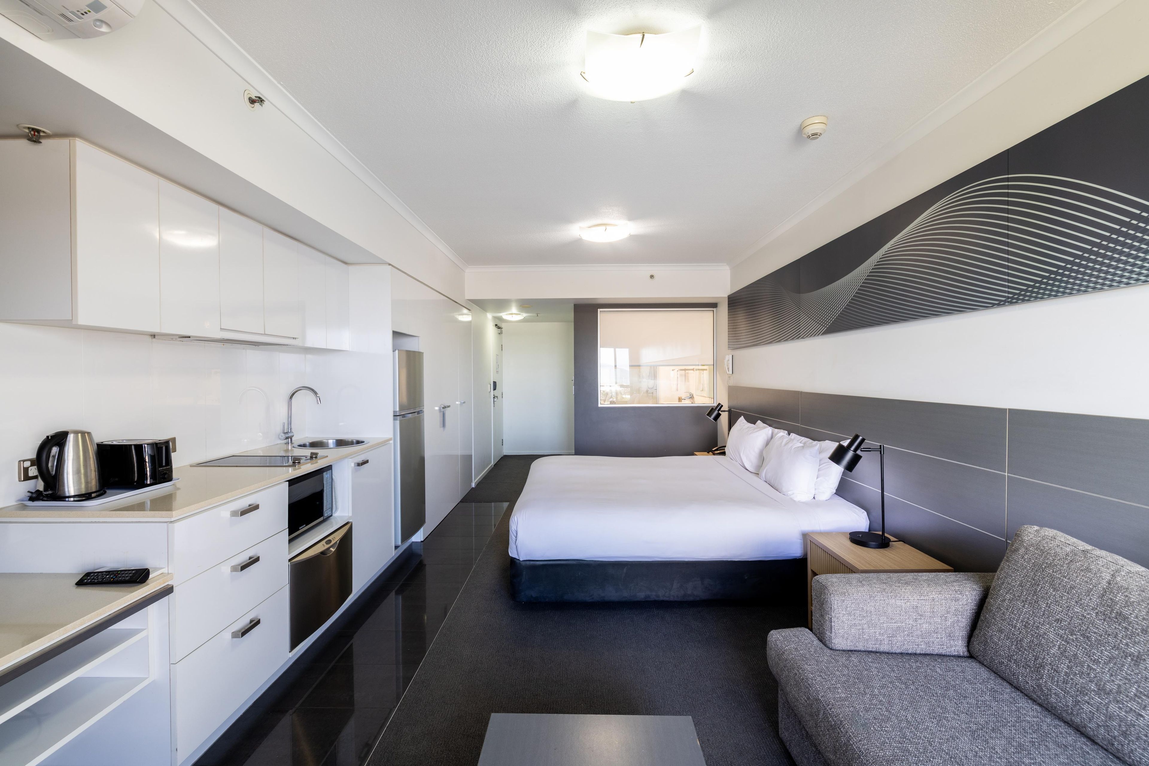 Una habitación de hotel moderna con kitchenette, una cama, un sofá y un ventanal grande.